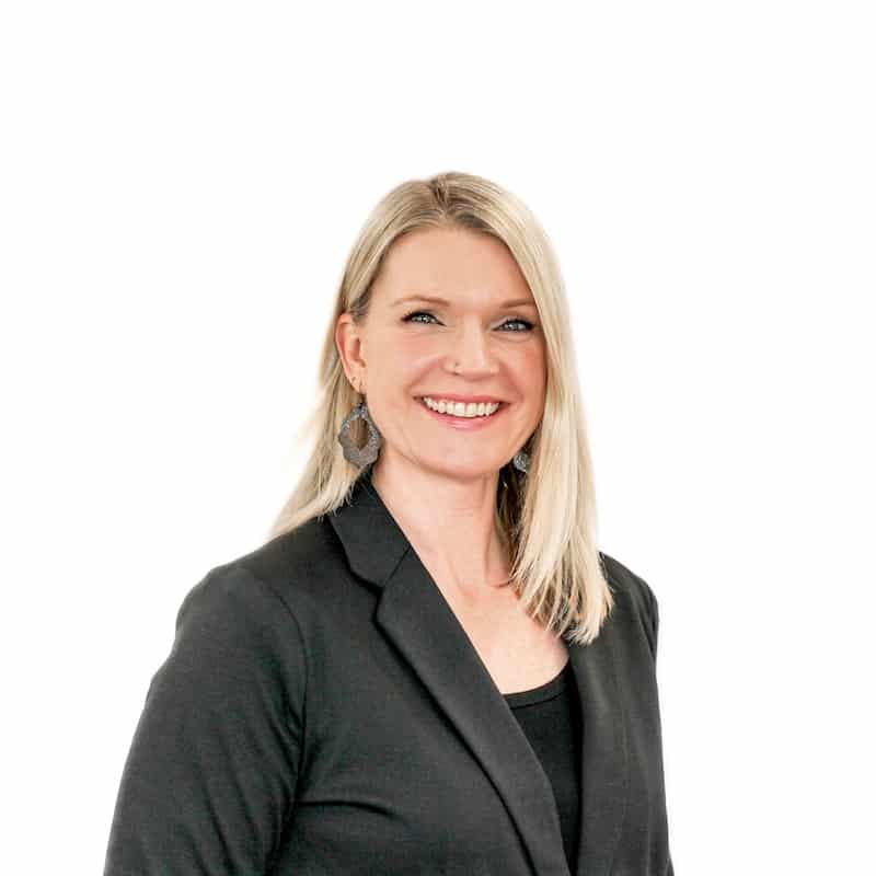 A smiling woman with blonde hair from a beauty salon in Omaha, wearing a black blazer against a white background.