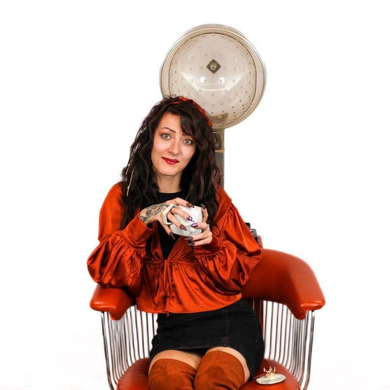Woman sitting in a retro beauty salon hair dryer chair holding a cup.