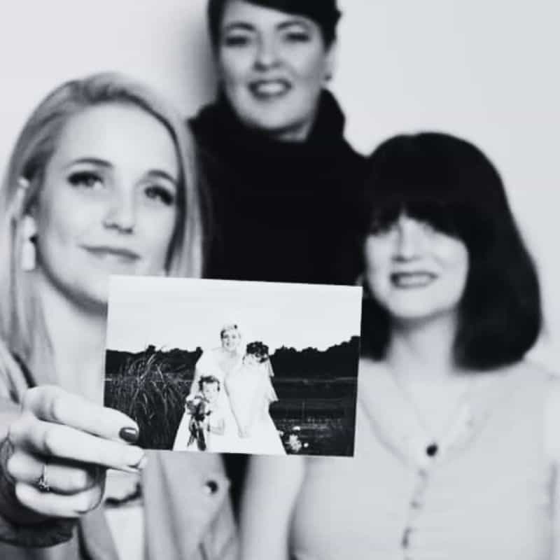 Three generations of women holding a family photograph outside a hair salon.