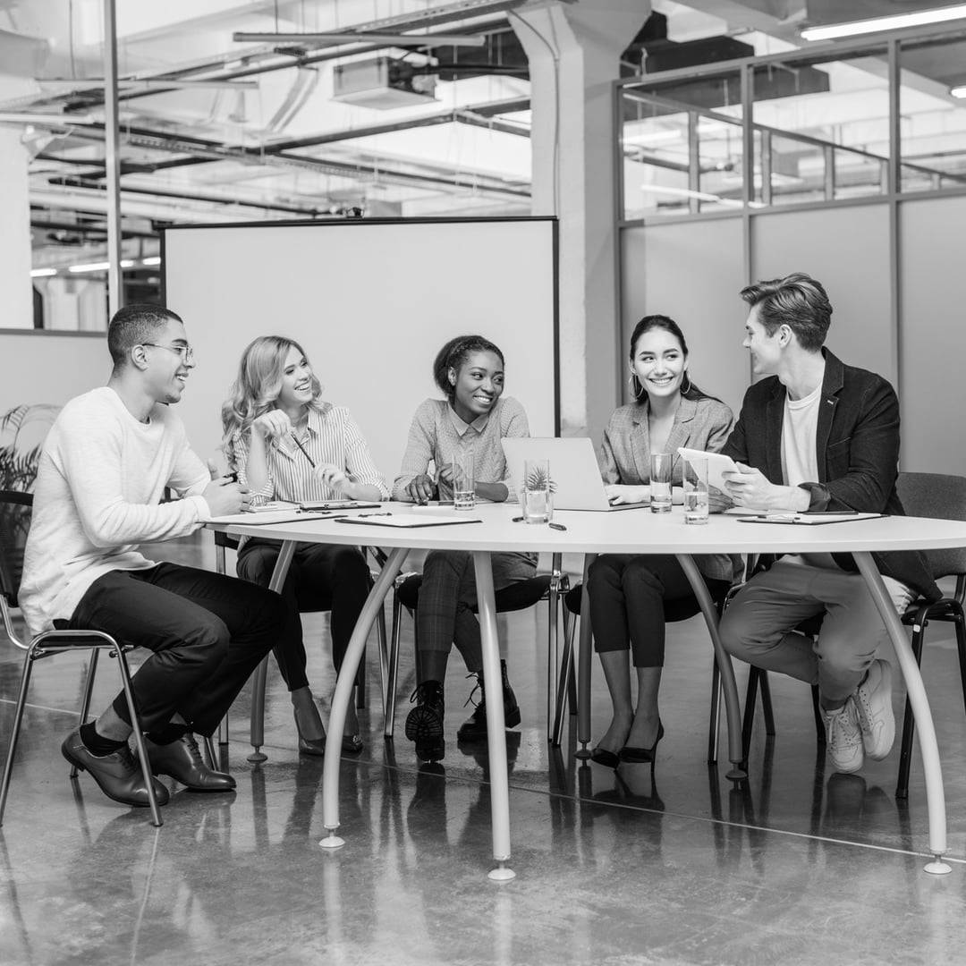 Five people sitting around a table in an office, engaged in a meeting and smiling.