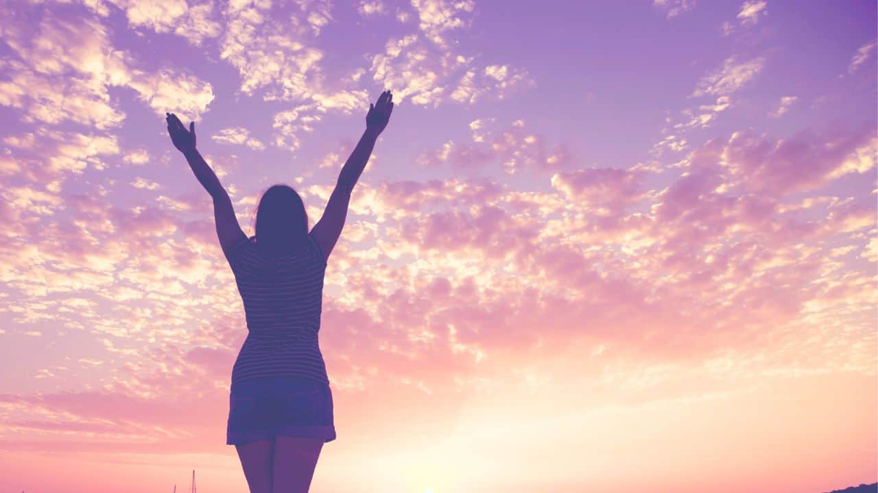 Woman with arms raised in triumph against a sunset sky, her hair styled exquisitely after a visit to the cosmetology school.