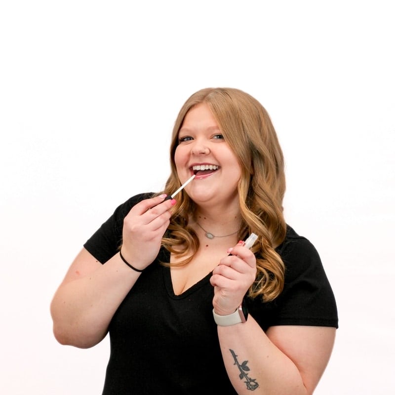 A smiling woman applying lip gloss at a beauty hair school.