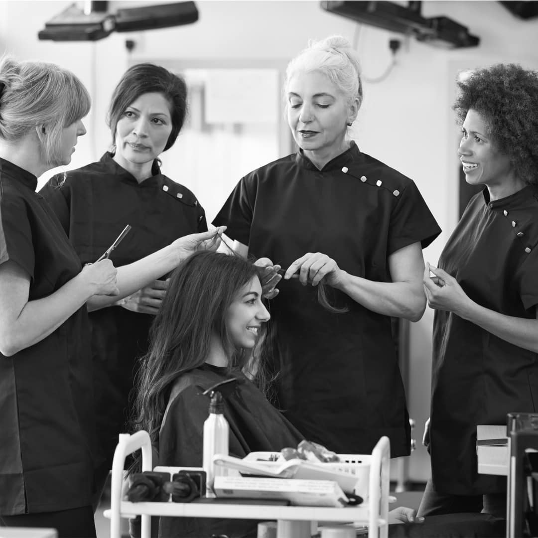 A group of hair stylists in black uniforms styles a seated woman's hair at a salon.