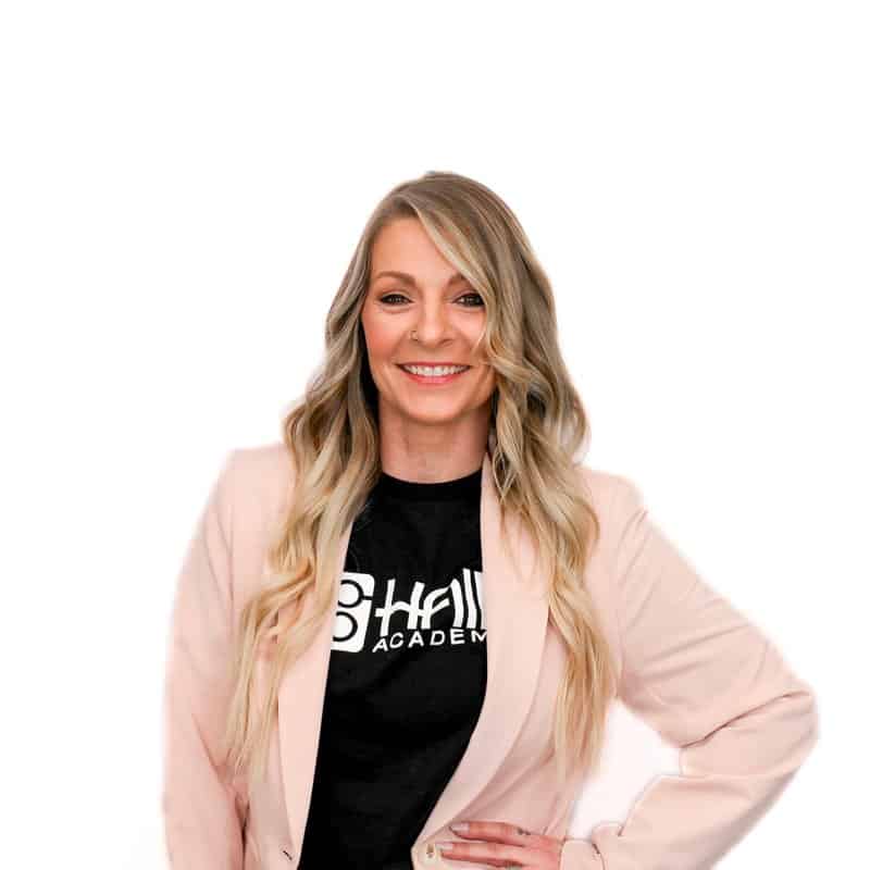 Woman smiling while standing with arms akimbo, wearing a pink blazer and a black t-shirt with white lettering, exuding the confidence of a cosmetology instructor.