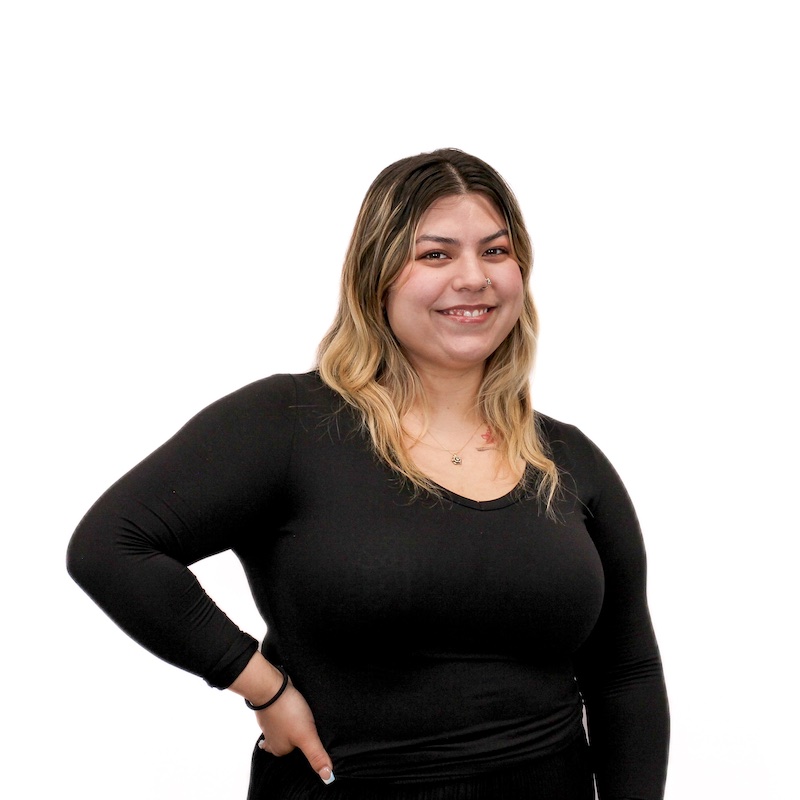 A smiling person with long, blonde hair wearing a black long-sleeve top, posing with one hand on their hip against a plain white background.
