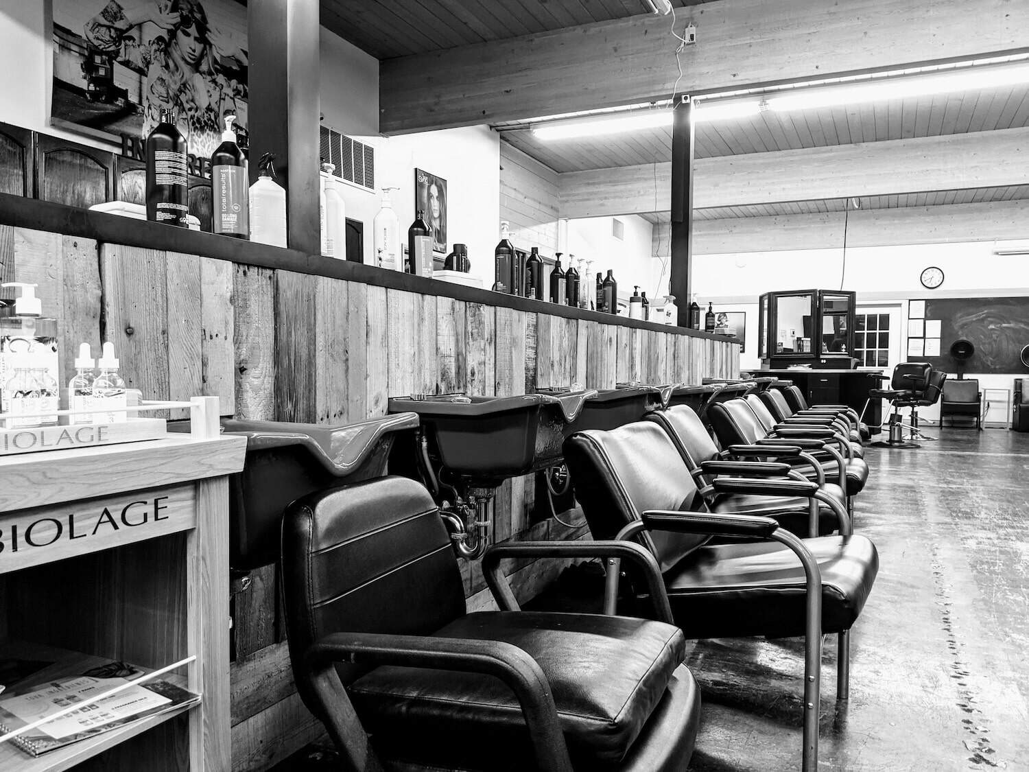 Black and white photo of an empty hair salon, showing a row of washing stations with chairs and shelves stocked with hair products.