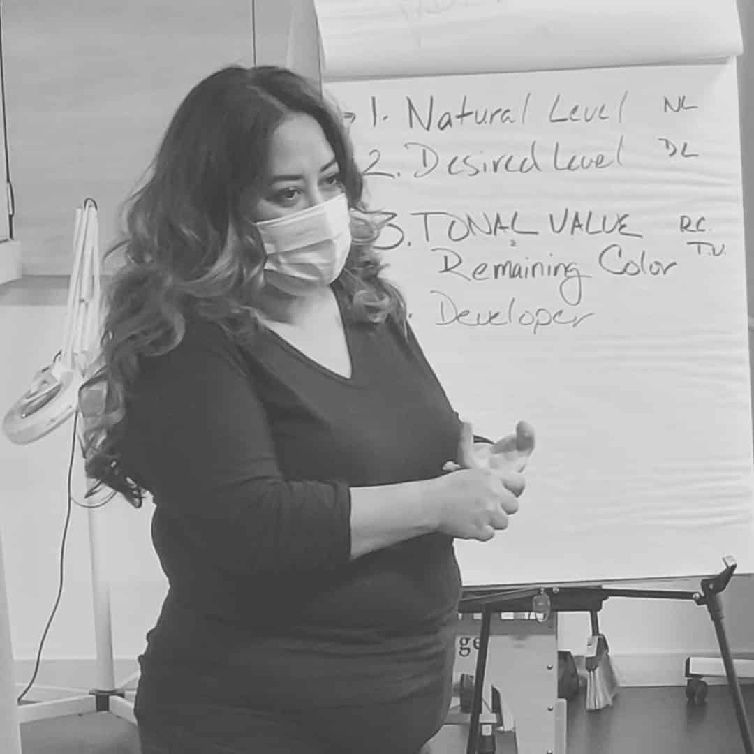 Person in a mask stands in front of a flip chart discussing hair color levels and tonal value.