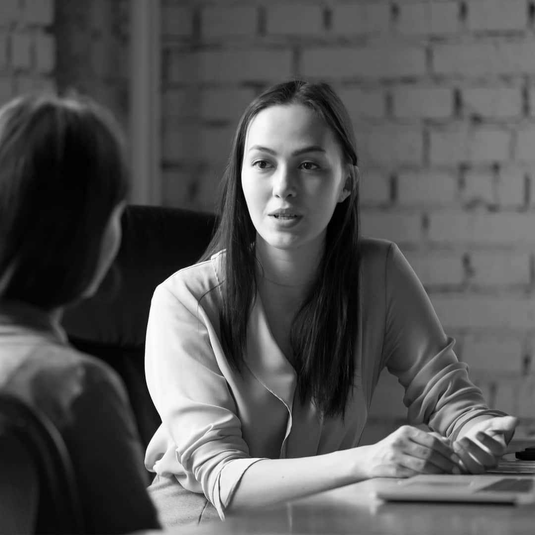 Two people sitting across from each other at a table, engaged in conversation, in a room with a brick wall backdrop. One person is gesturing with their hands. Black and white image.