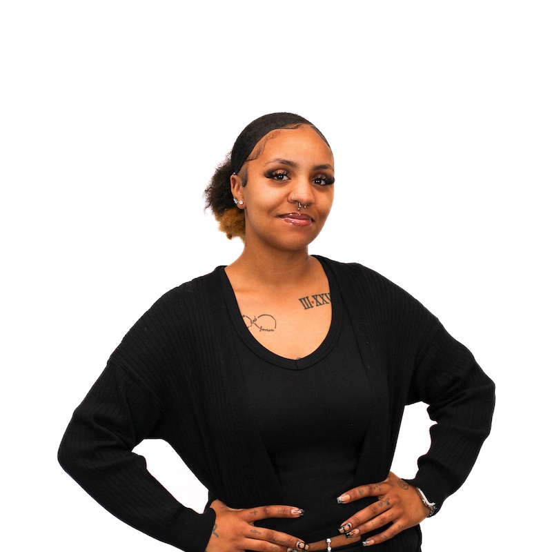 A person with a confident pose stands against a white background, wearing a black outfit and displaying tattoos and a nose ring.