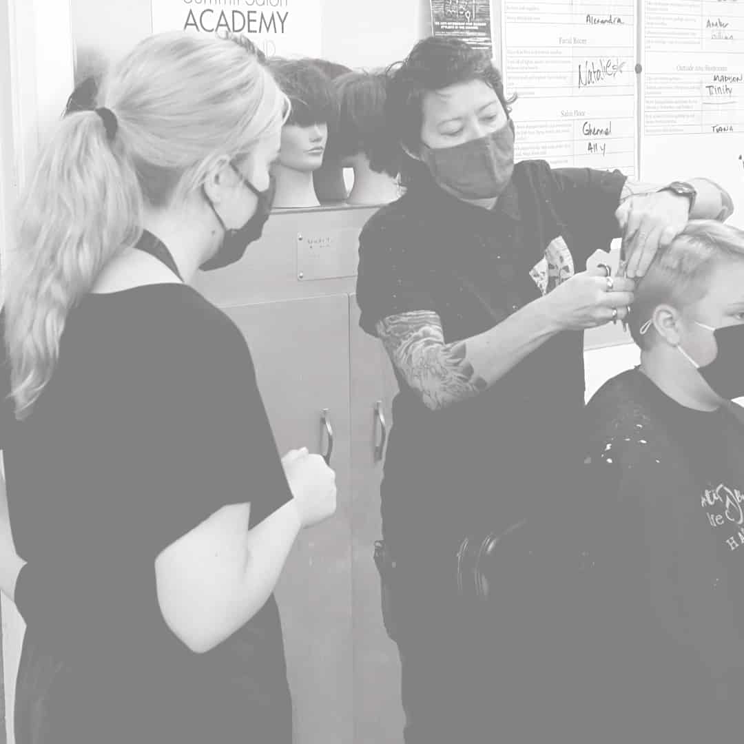 Stylist cutting a client's hair while another person observes, both wearing masks in a salon environment.
