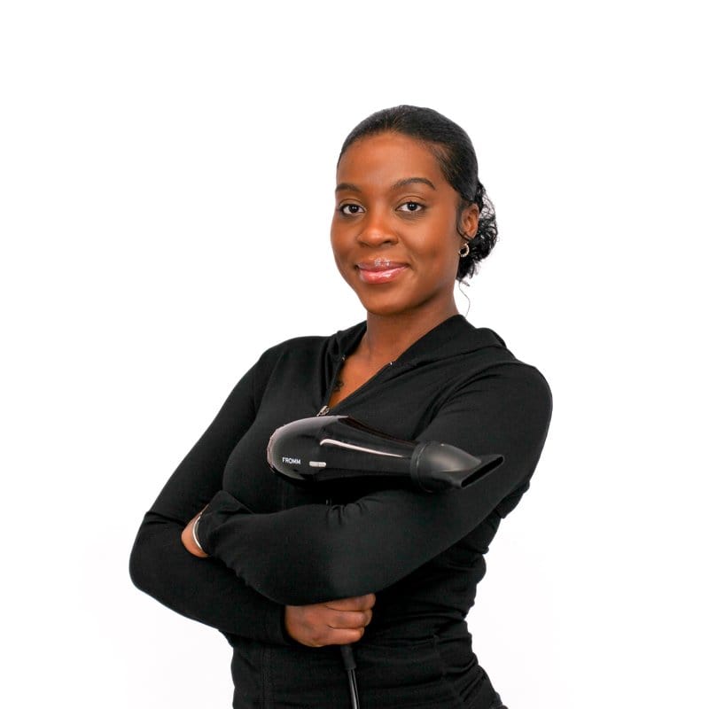 Woman with a confident expression standing with arms crossed while holding a hair dryer, showcasing her expertise in cosmetology.
