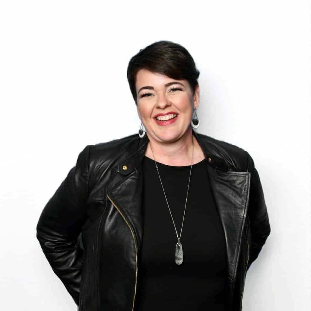 A woman with short dark hair styled from a cosmetology school smiling while posing with her hands on her hips, wearing a black leather jacket and black top against a white background.